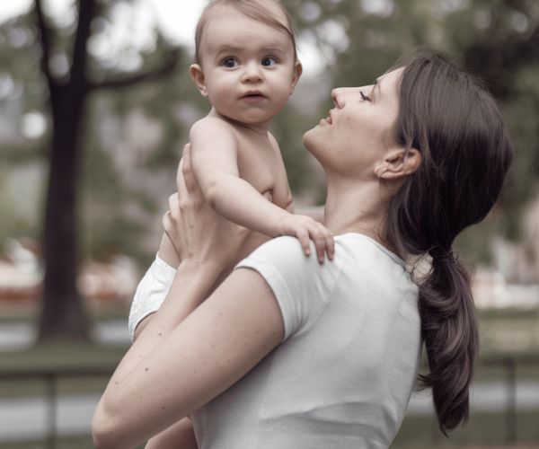 Mother with back pain in a park holding baby up in the air. Mother looking adoringly at her baby whilst baby looks away from Mother towards viewer.